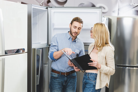 consultant pointing with hand at folder and talking with woman near fridges in home appliance store