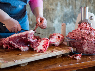 beautiful mature woman in pink sweater and gray economic apron cuts large piece of pork into pieces with knife and ax. girl puts small pieces in meat grinder and grinds them into minced meat