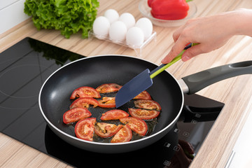 Slices of red tomatoes fried in oil on pan on stove. Woman's hand of young housewife holds culinary spatula and flips stewed vegetables for scrambled eggs. 