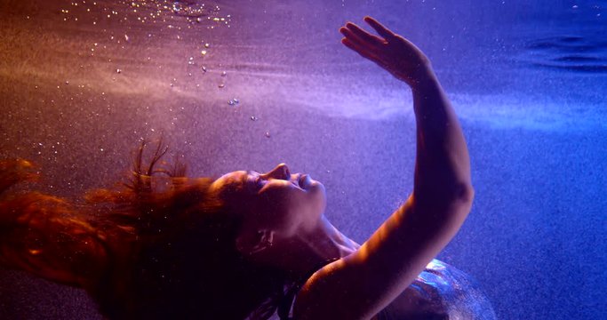 woman is sinking inside water of swimming pool, stretching hand to surface, underwater view