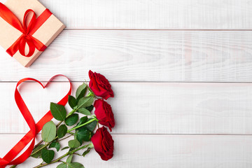 Valentines day romantic table setting, red tape in shape of heart, red fresh roses, kraft gift box with bow on white wooden background. Copyspace, empty place for text. Top view, flat lay, horizontal.