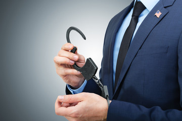 White man in suit holding handcuffs in hand with USA flag pin on his chest