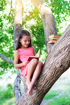 Charming little girl with long brown hair reads book outdoor sitting on tree in summer park or in a forest glade. Child booklover. Education, reading, kid's leisure