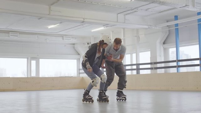Following shot of male professional inline skater teaching young woman wearing safety equipment and roller-skates. Woman falling down