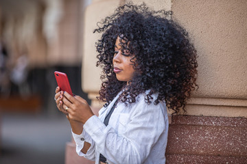 Young curly hair black woman walking using cell phone. Texting on street. Big city.