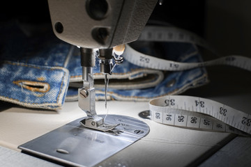 Needle of white industrial sewing machine use sew cloth with tape measure and blue jeans close-up.