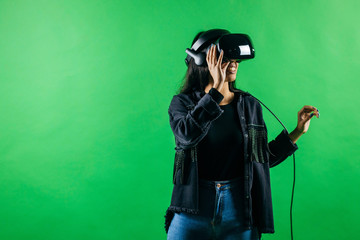 Mod curly dark haired girl dressed in black denim jacket uses the virtual reality glasses on her head and gloves in hands in the studio over green background