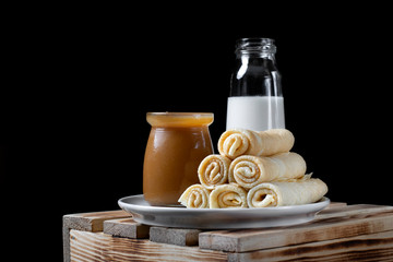 Fototapeta na wymiar Stack of rolled crepes on the ceramic plate, a glass jar of salted caramel and a bottle of milk