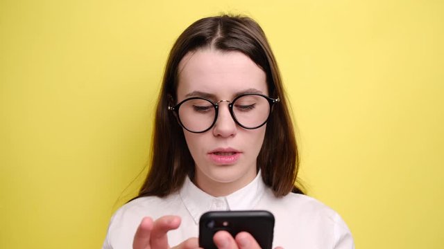 Shocked happy brunette woman in spectacles holds modern cell phone, stares at awesome invitation recieved via internet, reads impressive good news, isolated over yellow background