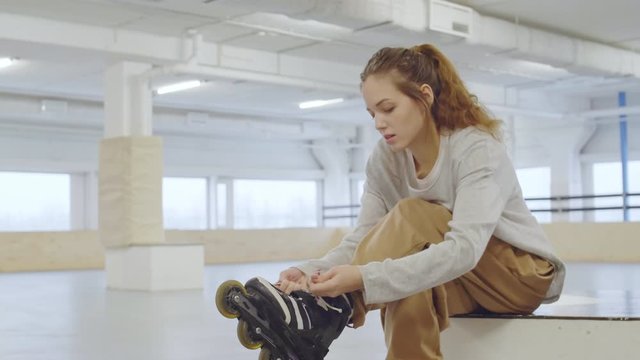 Lockdown of Caucasian woman with long curly hair putting on and lacing up roller-skates while sitting in indoor inline skating park