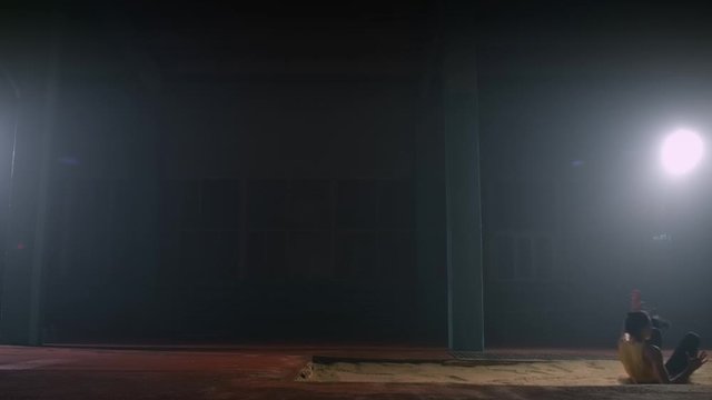 A Sporty Woman Runs Up In A Stadium And Performs A Long Jump In Slow Motion In A Stadium On A Dark Background