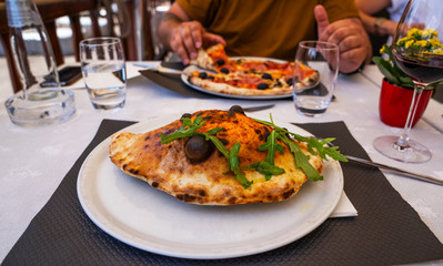 Baked calzone on the plate. Closed type of pizza, decorated with basil twigs on a restaurant table background.