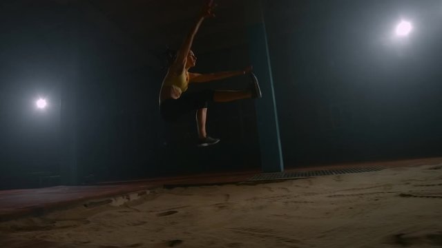 Running woman athletics at the stadium on a treadmill performs a triple jump in length and flies into the sand landing on her feet. Slow motion