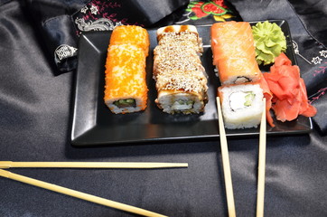 Sushi is a Japanese dish that consists of glutinous rice with fish, seafood and vegetables. On a black background is a plate with sushi, ginger, soy sauce and wasabi, wooden sticks.