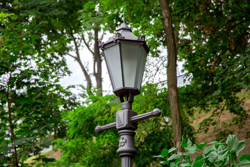 iron street lamp in retro style with glass inserts, in the park in the background trees with branches and green leaves on a summer day.