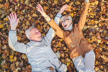 Cheerful active senior couple lying on ground rolling around in autumn leaves having fun. Positivity