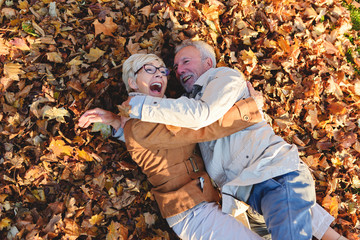 Cheerful active senior couple lying on ground rolling around in autumn leaves having fun. Positivity