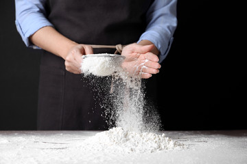 Woman sifting wheat flour at table against black background, closeup
