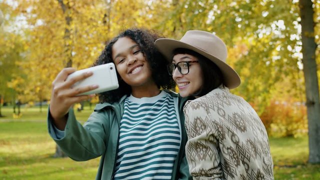 Attractive young women are busy taking selfie in park using smartphone camera posing having fun outdoors. Modern lifestyle, devices and nature concept.