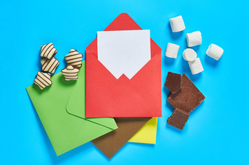 Scattered sweets, striped candies, cylindrical marshmallows, broken chocolate near envelope with white blank paper sheet lies on blue desk. Love letters concept. Close-up