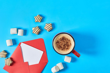Scattered striped candies, marshmallow near envelope with empty paper sheet and full cup of coffee lies on blue desk. Love letter concept. Space for text. Top view