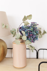 Blue and purple hydrangea in a beige vase stands on a wooden shelf on a white background, eco-friendly home decoration, flowers for march 8