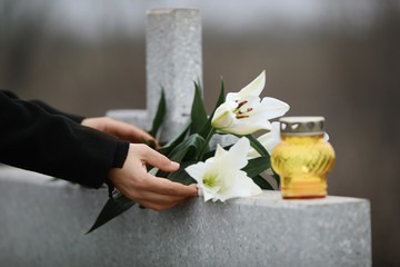 Woman holding white lilies near light grey granite tombstone with candle outdoors, closeup. Funeral...