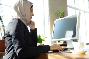 Looking at screen. Portrait of a beautiful arabian businesswoman wearing hijab while working at openspace or office. Concept of occupation, freedom in business, leadership, success, modern solution.