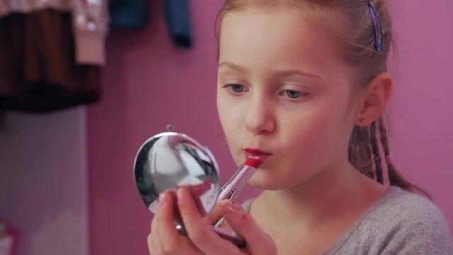 Little girl paint lips with red lipstick looking in mirror in childrens room nursery white furniture pink wall on background closeup