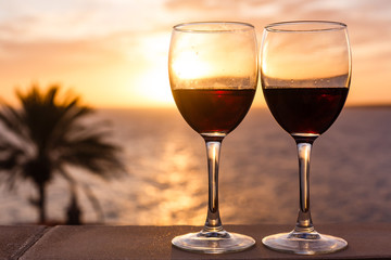 Two glasses of red wine sitting on a ledge over looking the beach, ocean and a beautiful sunset.