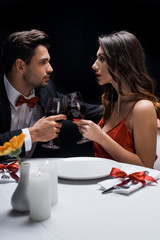 Side view of elegant couple looking at each other while clinking with wine at served table isolated on black