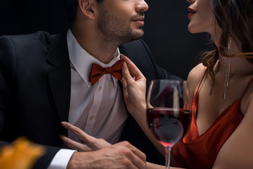 Cropped view of elegant woman adjusting bow tie of man in suit by wine glass isolated on black