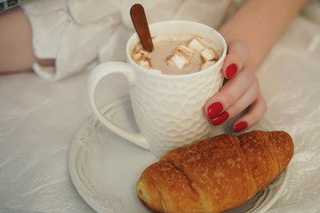 A young woman eats a croissant for Breakfast and drinks coffee. A woman's hand holds a mug of coffee on a plate is a croissant
