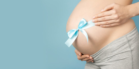 Beautiful young pregnant woman holds hands on her belly on blue background. Motherhood, pregnancy, expectation of the child, happiness concept. Ribbon on pregnant belly. Close-up, place for text