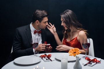 Side view of handsome man presenting jewelry ring at smiling girlfriend during romantic dinner isolated on black