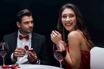 Smiling woman looking away by handsome man with jewelry ring at served table isolated on black