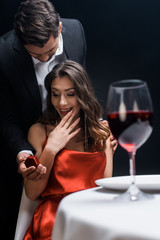 Handsome man presenting box with jewelry at exited woman during romantic dinner isolated on black