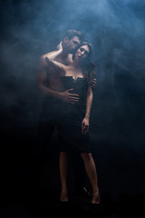 Full length of muscular man kissing sexy woman on black background with smoke
