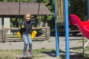 little girl child in a motorcycle jacket rides a swing and shows a thumb on her hand