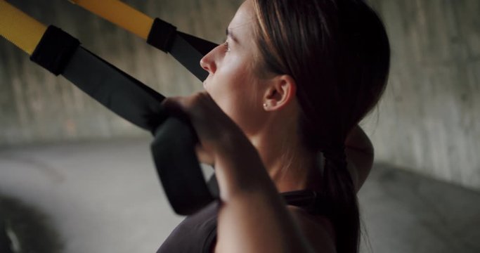 Side close up shot of concentrated powerful female using TRX loops sport lifestyle, motivation of perspiring Caucasian girl breathing doing pull ups exercise training for arms muscles outdoor urban