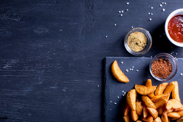 Baked potato wedges in a pan, spices and red sauce on a black wooden background. Background menu food.