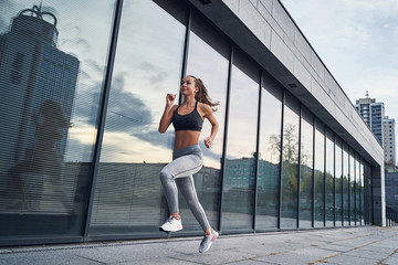 Young athletic woman running in the city, shot of girl runner on the glass building background