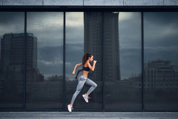 Young athletic woman jumping while making running training at urban city location with big mirror windows on background