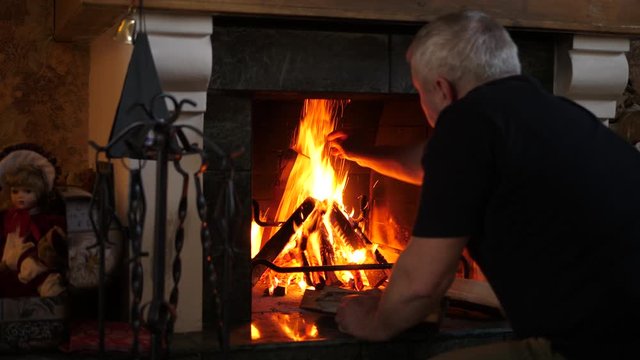Old man adding more fire wood to fireplace at home in the attic. Evening time.