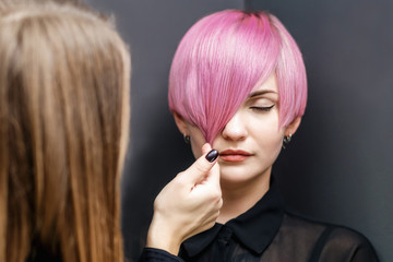 Hairdresser checking short pink hairstyle of young woman on gray background.
