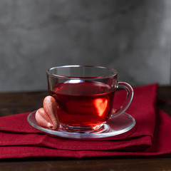 A glass cup of red tea Hibiscus and meringue cookies in the shape of hearts. Stand on textile napkin, wooden table with a concrete background. Still life dark photo minimalism with copy space.