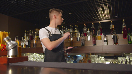 Bartender stands with tablet and counts bottles with alcohol