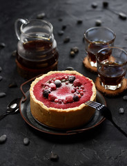 Homemade comfort food. Open lingonberry pie with raw raspberries and currants in metal tray with black coffee in glasses on black background