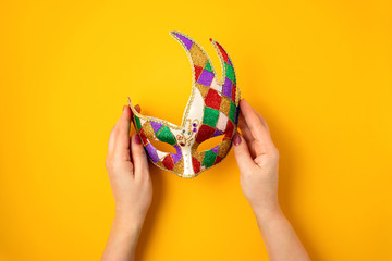 Fototapeta premium Woman hand holding festive, colorful mardi gras or carnivale mask over yellow background. Party invitation, greeting card, venetian carnivale celebration concept. Flat lay, top view