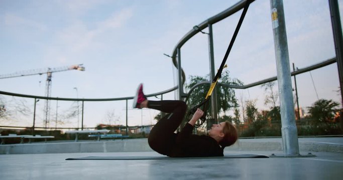 Side shot of purposeful young female lye down on sport mat using TRX suspension straps, outdoor urban sportive yard spot for healthy success fitness workout exercising woman instructor in black